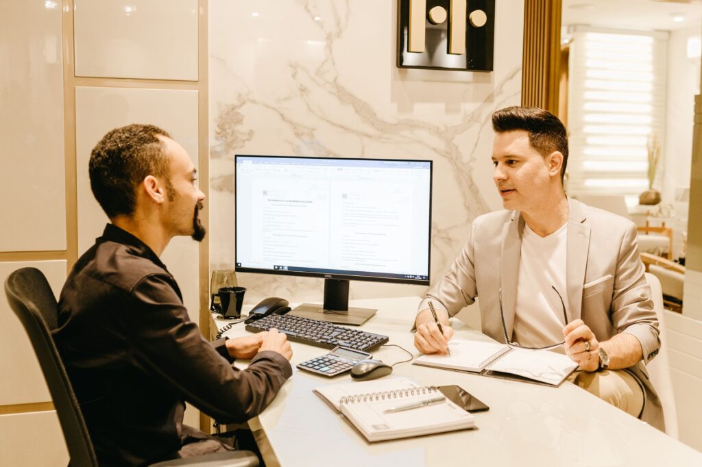 Two businessmen discussing work at a sleek and modern office desk, using computer and notes for collaboration.